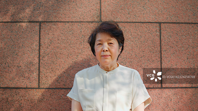 一位老年女性站在石墙前，开心地看着镜头并微笑。图片素材