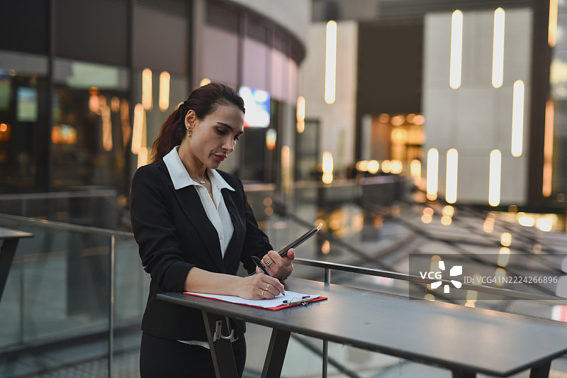 微笑的女商人在办公室阳台上站着写笔记。图片素材