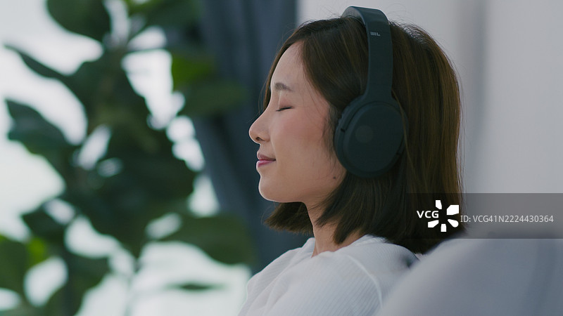 放松的年轻亚洲女性在家中戴着耳机享受音乐以提升身心健康。图片素材
