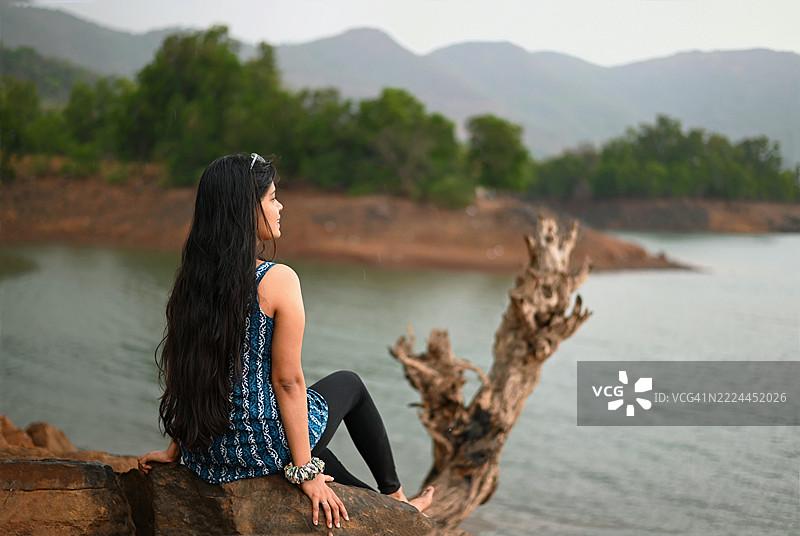 年轻女子坐在锈色悬崖岩石上，俯瞰宁静的碧绿色水库图片素材