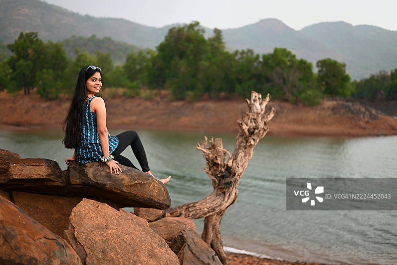 年轻女性坐在锈色的悬崖岩石上，微笑着看向镜头图片素材