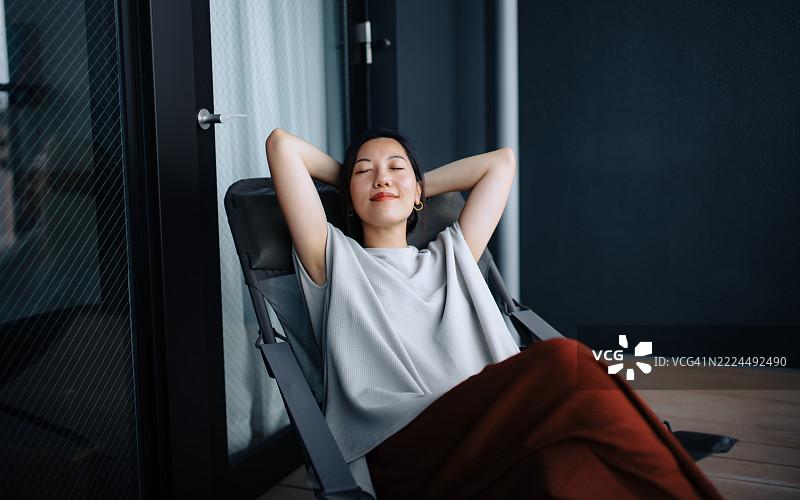 无忧无虑的年轻亚洲女性坐在阳台的躺椅上，闭着眼睛，双手放在头后，享受着晨光和宁静。心理健康与幸福感。健康与正念的概念。图片素材