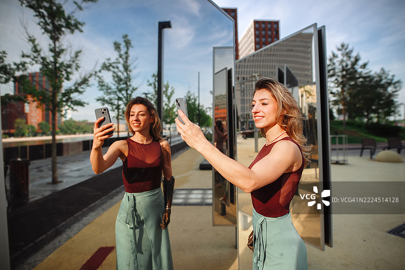公园的夏日氛围：一位穿裙子的瘦女人在镜子旁跳舞，感受着自信与快乐，正在用智能手机拍摄生动的自拍。图片素材