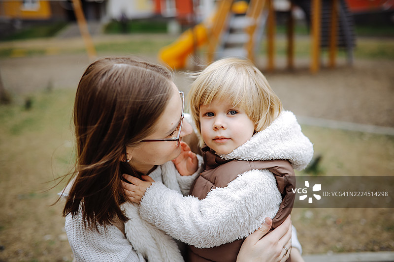 年轻家庭在新住宅小区，母亲在游乐场拥抱她的幼儿儿子。图片素材