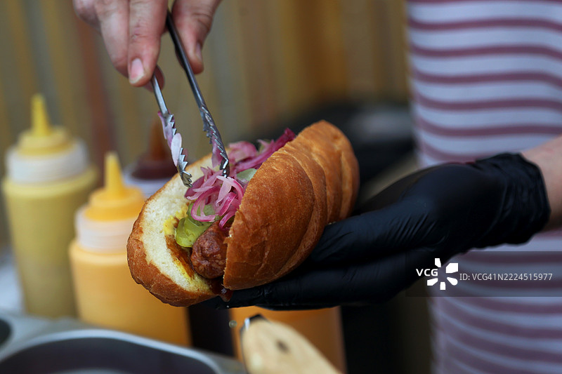 厨师的手在户外小吃摊制作热狗，配有香肠、红洋葱、腌黄瓜和芥末酱。图片素材