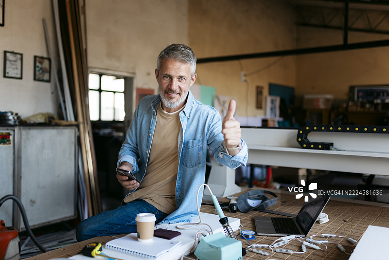 微笑的工程师在车间使用智能手机并竖起大拇指。图片素材