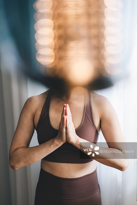 一位女性站在窗边，双手合十祈祷。自然光下的宁静正念练习。图片素材