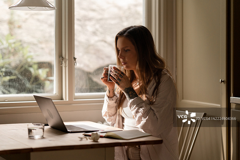 在舒适的厨房里，一位女性在家工作，享受着咖啡，旁边放着一台笔记本电脑。图片素材