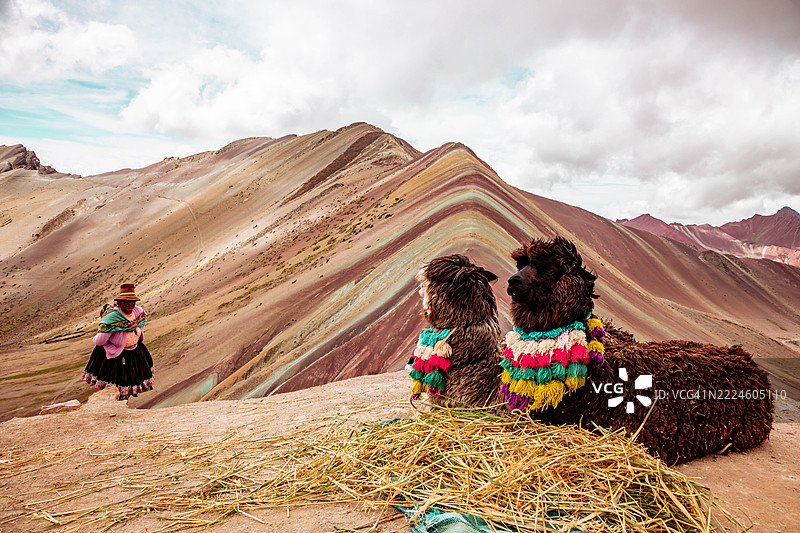 前景中有两只羊驼，背景是秘鲁的维尼昆卡彩虹山和当地农村女性。图片素材