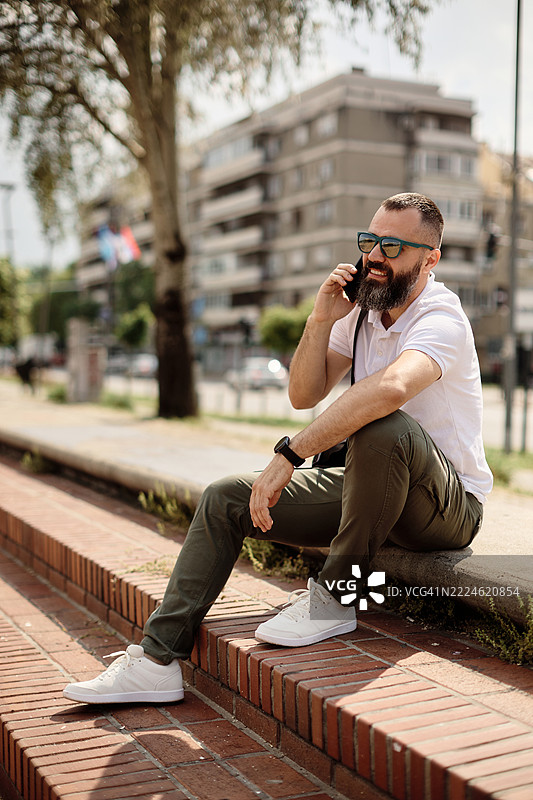 一位年轻帅气的有胡子的男人坐在码头上打电话。图片素材