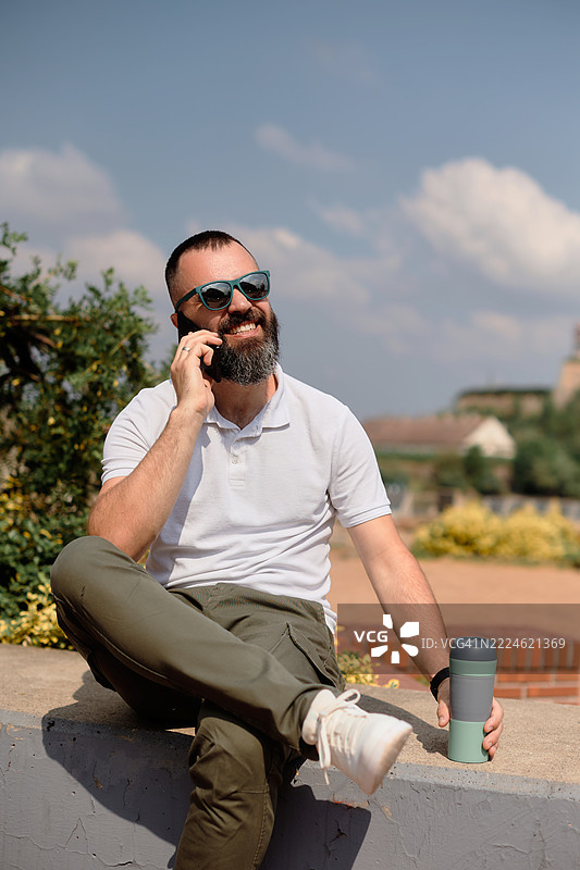 一位年轻帅气的有胡子的男人坐在码头上打电话。图片素材