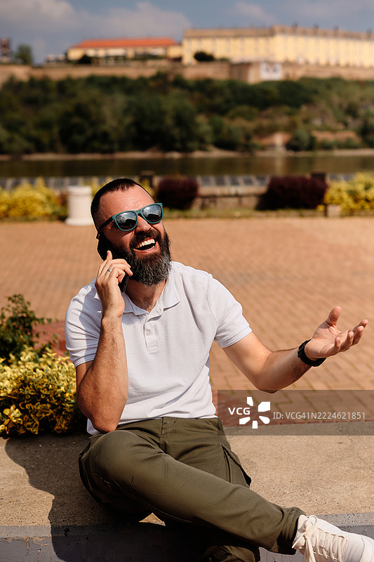 一位年轻帅气的有胡子的男人坐在码头上打电话。图片素材