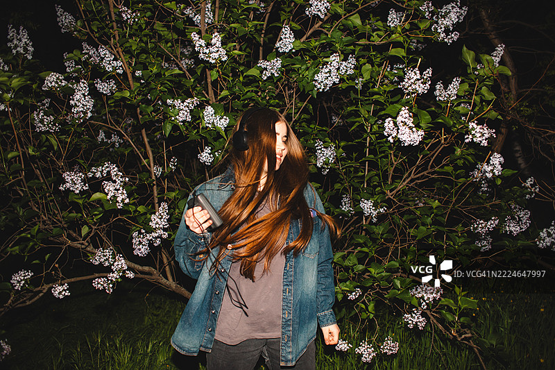 快乐的年轻女性在紫丁香花丛中一边听着耳机里的音乐，一边手持个人卡带播放器跳舞。图片素材