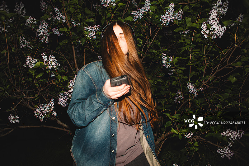 年轻女性在紫丁香花丛中一边跳舞一边戴着耳机听音乐，手里拿着个人卡带播放器。图片素材
