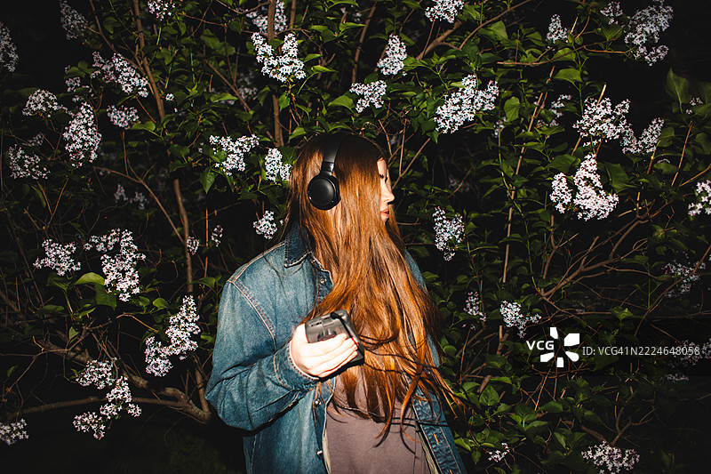 年轻女性在紫丁香花丛中戴着耳机，手持个人卡带播放器听音乐图片素材