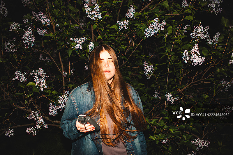 年轻女性在紫丁香花丛中戴着耳机听音乐，手中拿着个人卡带播放器图片素材