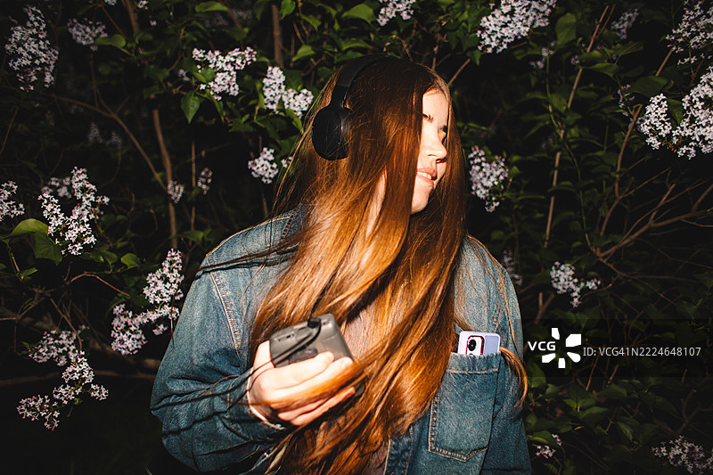 快乐的年轻女性在紫丁香花丛中一边听着耳机里的音乐，一边手持个人卡带播放器跳舞。图片素材