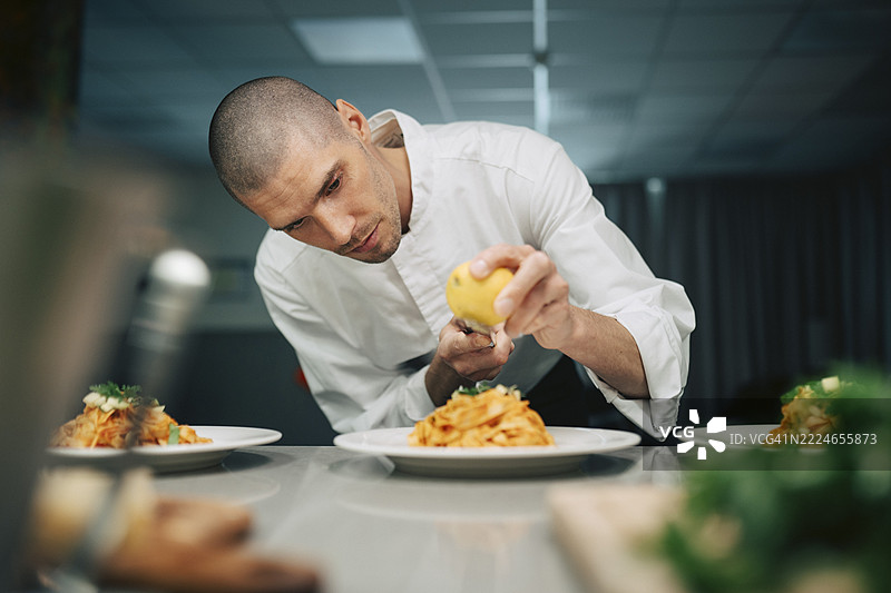 专注的男性厨师，光头，在餐厅厨房为意大利宽面条装饰柠檬皮屑图片素材