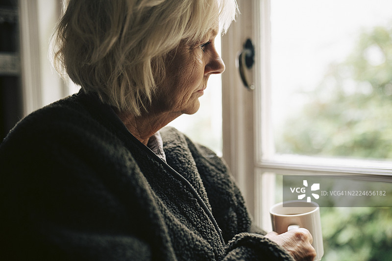沉思的老年女性手握咖啡杯，望着家中的窗外。图片素材