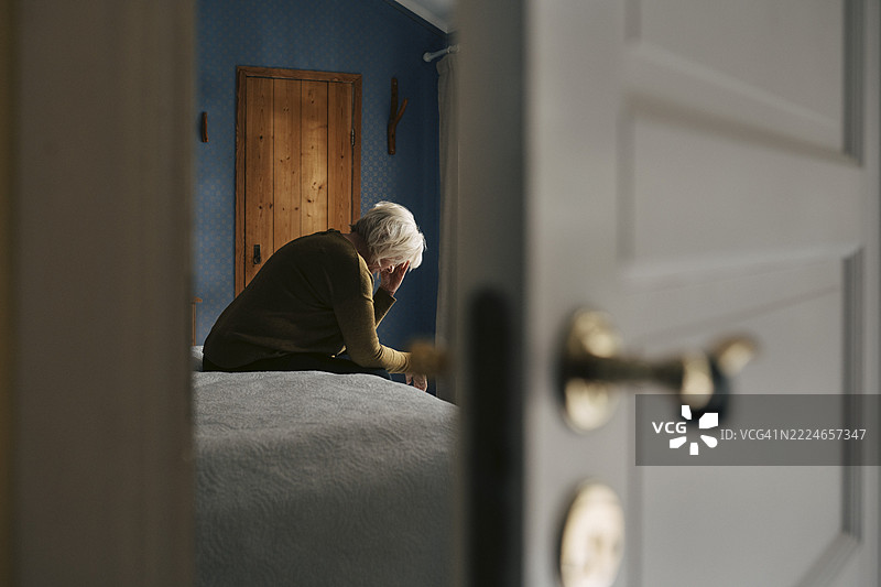沮丧的老年女性坐在床上，脸朝下，从卧室门口看到。图片素材