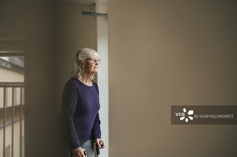在养老院靠近窗户站着的思考中的老年女性，手持拐杖。图片素材
