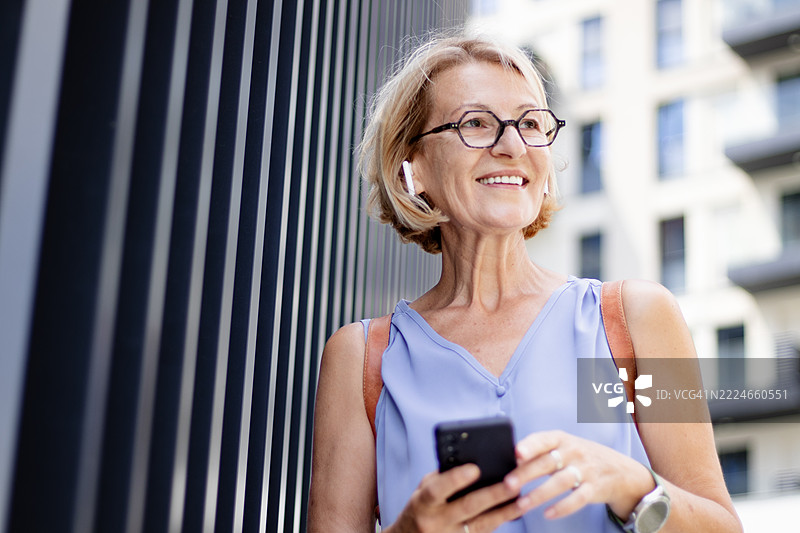 一位成熟的金发女性正在游览欧洲城市，佩戴无线耳机，边走边使用手机导航。图片素材