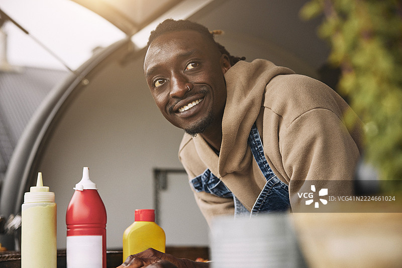 低角度视图的微笑男性餐车老板倚靠在柜台上，目光投向一旁。图片素材