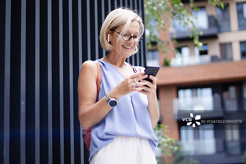 成熟的金发女性游客一边走在图表上，一边手持手机，佩戴无线耳机，使用移动应用程序。图片素材