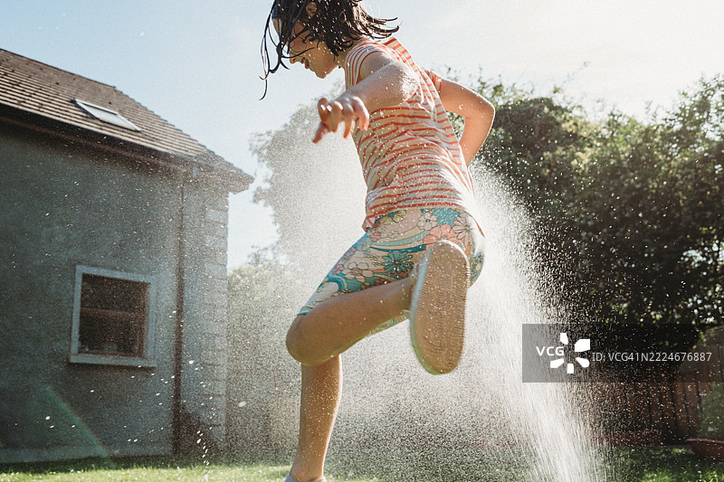 一个无忧无虑的小女孩在炎热的日子里，享受着在花园里通过喷头奔跑的清凉时刻。图片素材