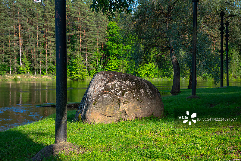 一块大型苔藓覆盖的岩石坐落在平静的河边绿草上，周围环绕着树木和高高的黑色路灯，拉脱维亚图片素材