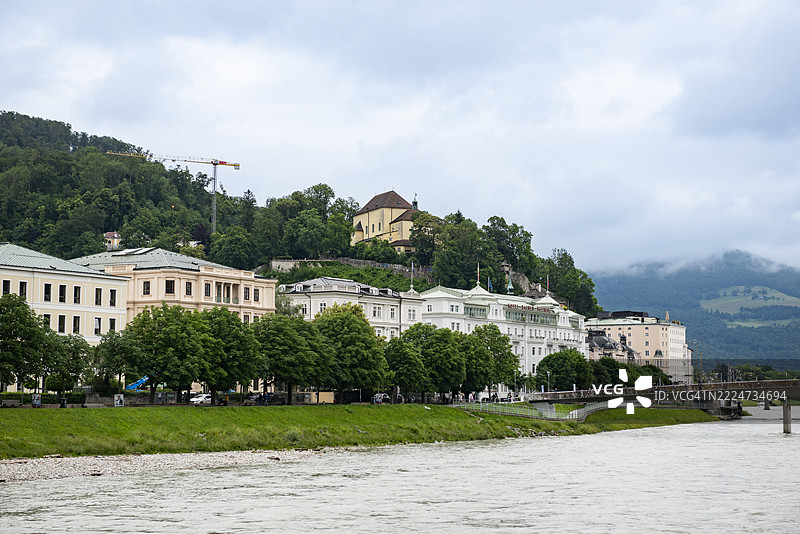 萨尔茨堡的河边景观展示了历史建筑、萨赫酒店和卡普齐纳山的森林坡道，天空阴云密布，位于奥地利萨尔茨堡。图片素材
