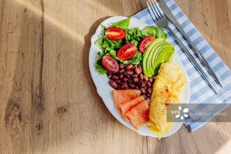 美味的早餐 - 木桌背景上的煎蛋卷、三文鱼、豆类、沙拉和牛油果。正上方，留有空白空间。图片素材