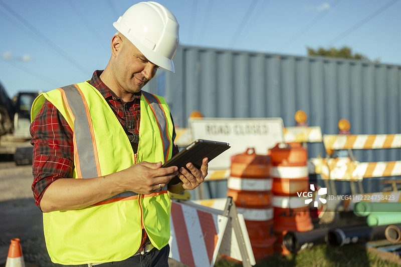 一名建筑工人在检查道路施工工作。他穿着白色安全帽和高可见度背心，这是建筑工地必备的防护装备。图片素材