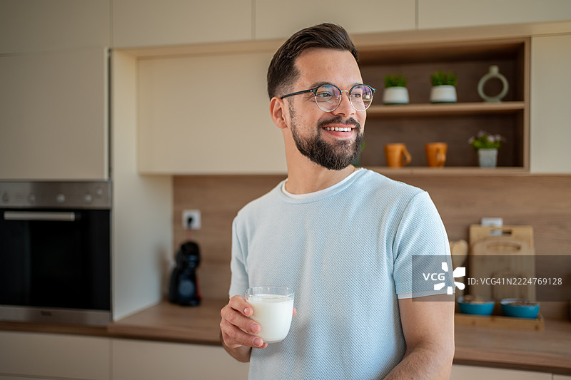 年轻男子在现代厨房里微笑着，手里拿着一杯牛奶图片素材