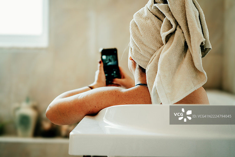 女人在浴缸中放松，头上裹着毛巾，使用智能手机图片素材
