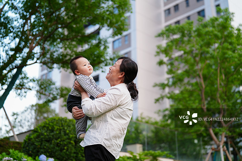 亚洲祖母微笑着抱着孙子，背景是有植物和建筑的住宅景观图片素材