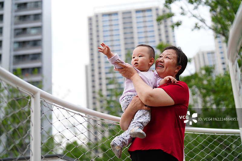亚洲祖母微笑着抱着孙子，背景是有植物和建筑的住宅景观图片素材