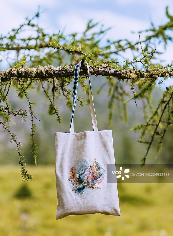 自然环境中悬挂在树枝上的帆布手提袋。环保帆布手提袋上绣有山脉插图的自然艺术作品，悬挂在阳光明媚的森林草地上的树枝上。图片素材