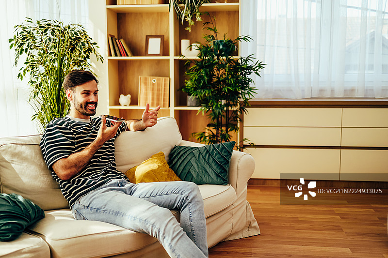 年轻男子坐在沙发上，使用手机的扬声器模式在家中。图片素材