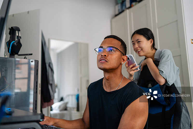 年轻男子在家工作，抬头看着身后微笑并拿着智能手机的女性。图片素材