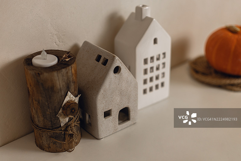 乡村风格的原木上放着一支蜡烛，旁边有两个房子形状的灯具，模糊的南瓜放在编织垫上。图片素材