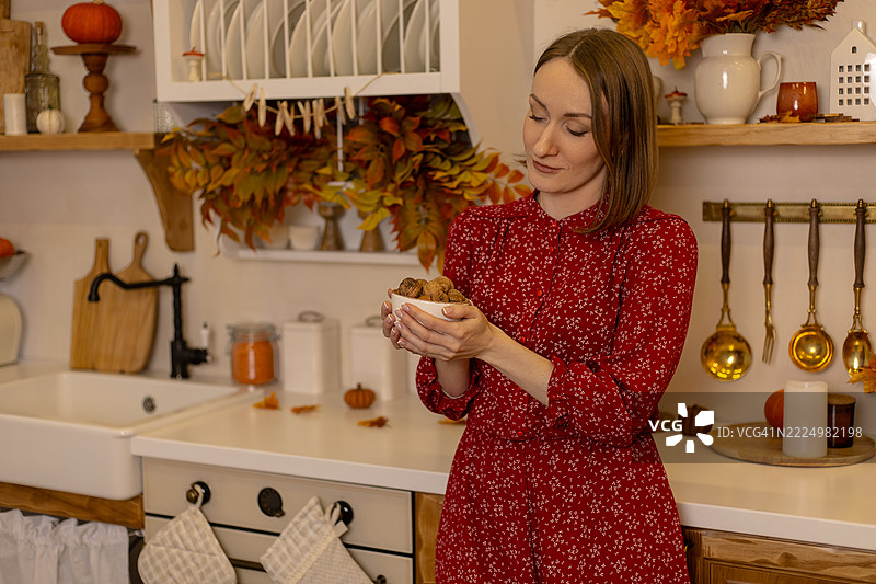 年轻女子穿着深红色连衣裙，在秋季温馨的厨房里，靠在台面旁，手里拿着一碗核桃。图片素材
