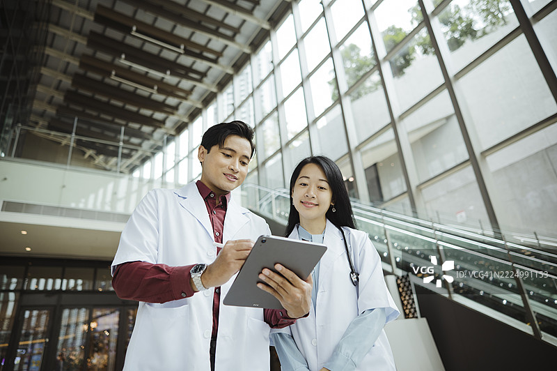 两位亚洲医生在现代医院里边走边谈，手持数字平板电脑。图片素材