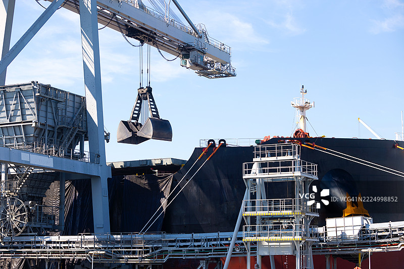 工业起重机在繁忙的港口将散装物料（如煤炭）从停靠的货船上吊起。在阳光明媚的港口进行物流和海运运输作业。图片素材
