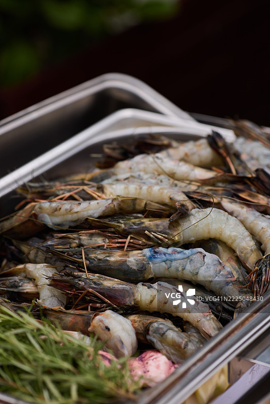 丰满的虾摆放在不锈钢托盘上，周围环绕着新鲜的香草，准备在烧烤派对上烤制。图片素材