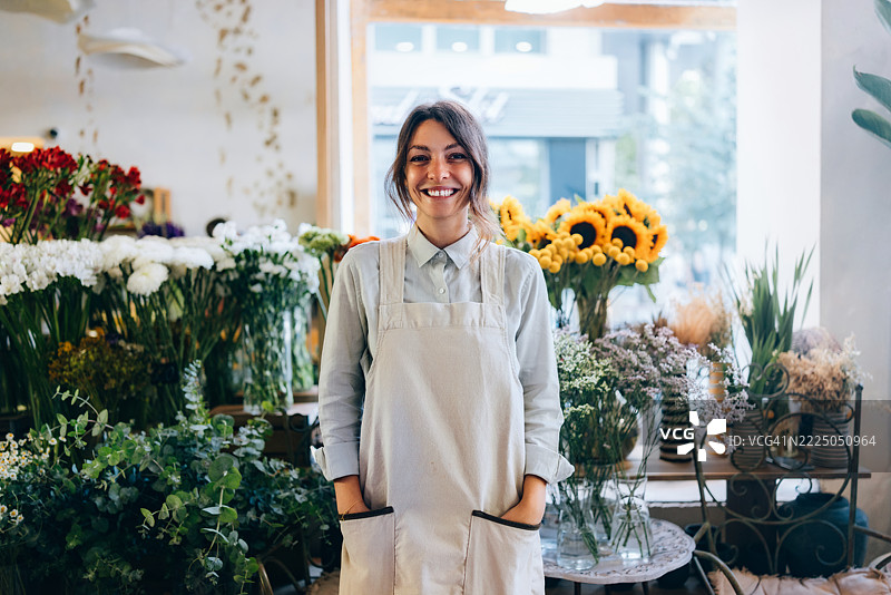 微笑的花店老板站在花店里，旁边是盛开的花卉布置图片素材