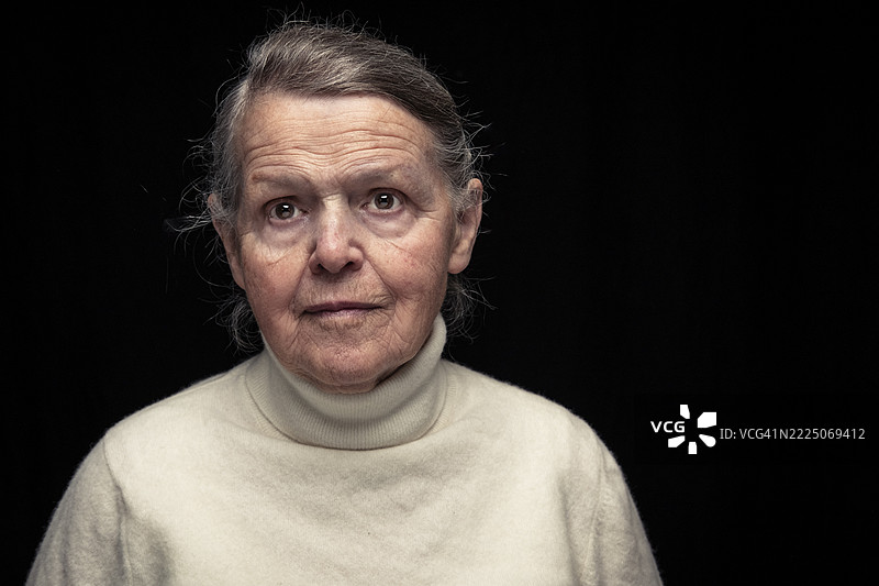 Elderly woman with blank expression standing against black background图片素材
