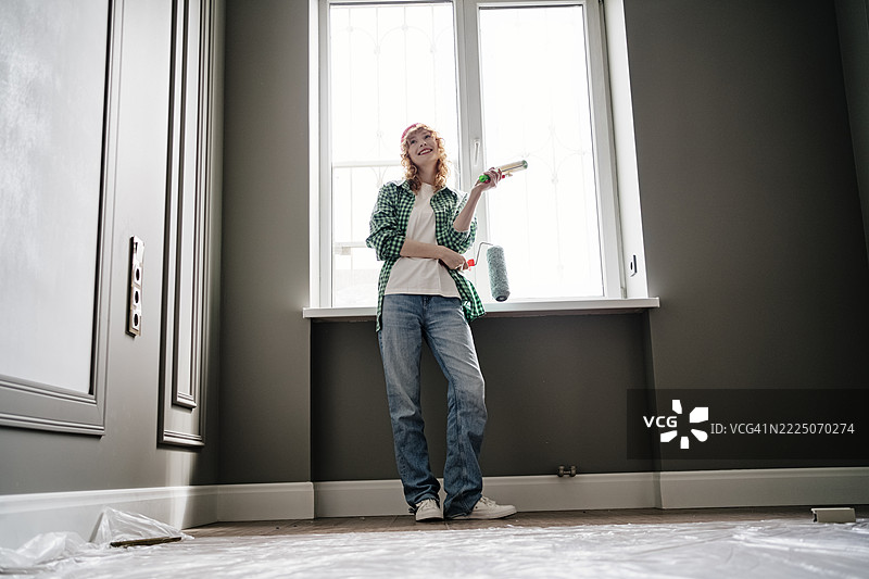 Happy young woman holding paint rollers in front of window at home图片素材