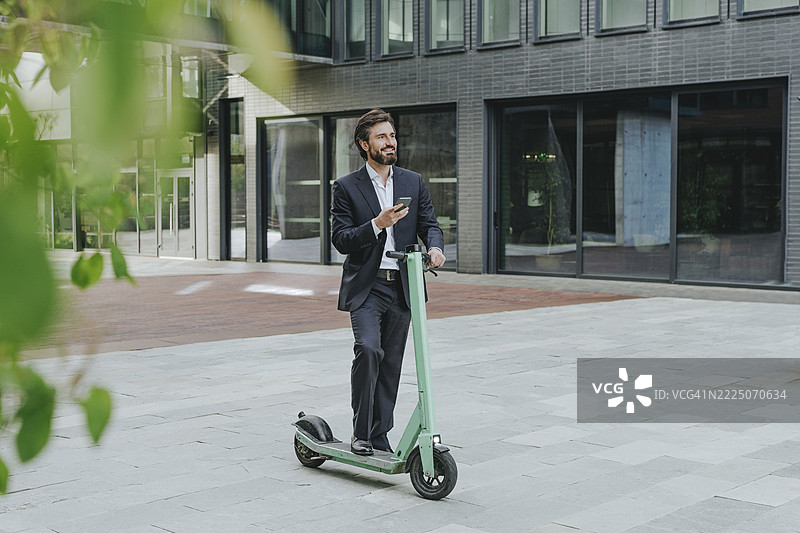Thoughtful businessman standing with electric scooter and holding smart phone at office park图片素材