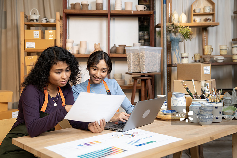 快乐的商业团队在陶艺店一起使用笔记本电脑。团队成员在陶艺工厂欣赏他们的数字艺术作品。小型企业图片素材